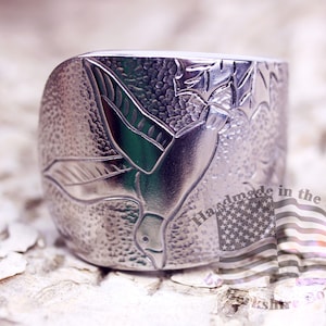 May include: A silver ring with a detailed engraved design of a bird in flight. The ring is handmade in the USA.