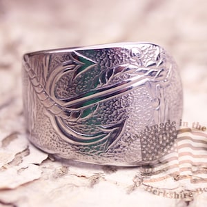 May include: A wide, silver ring with an engraved design of a fish hook and foliage. The ring has a textured surface and is likely made of metal. The words "handmade in the USA" are visible.