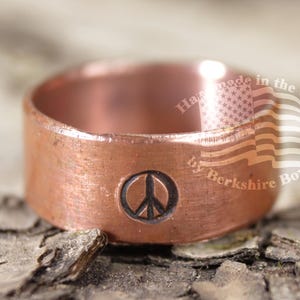 May include: A copper ring featuring a peace symbol. The ring has a wide band and a brushed finish. The text "Hand made in the USA by Berkshire Bowls" is visible on the side of the ring.
