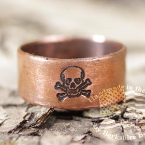 May include: A copper ring with a skull and crossbones design. The ring is wide and has a rustic, handmade appearance. The skull and crossbones are etched into the copper. The ring is displayed on a textured, wooden surface.