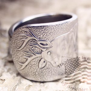 May include: A silver ring with a textured surface and a deer head design. The ring features an embossed deer head with antlers. The ring is marked "Handmade in the USA by Berkshire Bows."