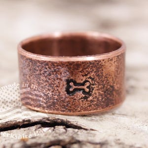 May include: A copper ring with a textured finish and a bone-shaped design engraved on the top.
