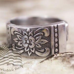 May include: A silver ring with a floral design. The ring is wide and has a textured surface. The design is intricate and detailed.
