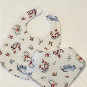 Op de afbeelding: Twee witte baby-slabbetjes met een Winnie de Poeh en Tigger print. De slabbetjes zijn gemaakt van zacht, absorberend materiaal en hebben een leuk, speels ontwerp.