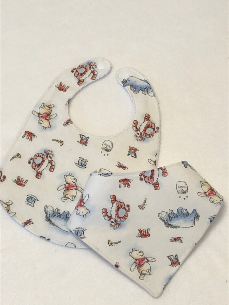 Op de afbeelding: Twee witte baby-slabbetjes met een Winnie de Poeh en Tigger print. De slabbetjes zijn gemaakt van zacht, absorberend materiaal en hebben een leuk, speels ontwerp.