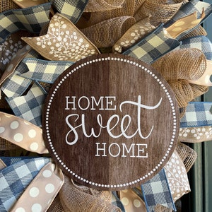 K&ouml;nnte beinhalten: Ein braunes Holzschild mit dem Text "Home Sweet Home" in wei&szlig;en Buchstaben. Das Schild ist von einem Kranz aus Sackleinen, blau-wei&szlig;em kariertem Band und braunem gepunktetem Band umgeben.
