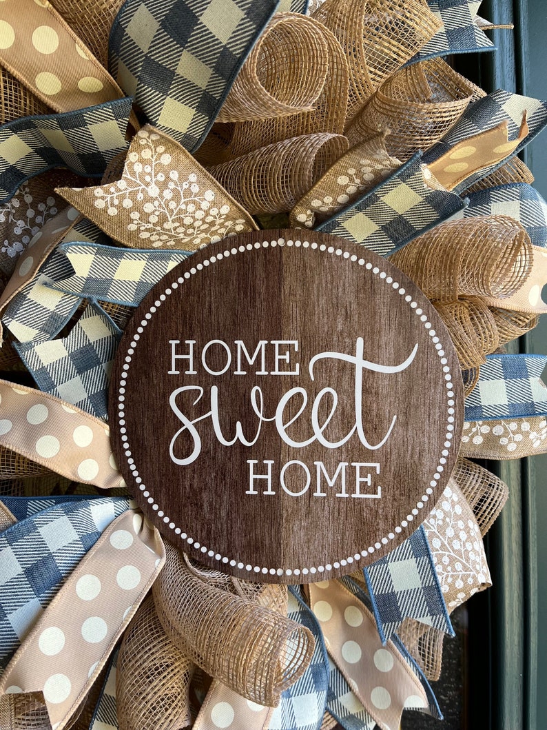 K&ouml;nnte beinhalten: Ein braunes Holzschild mit dem Text "Home Sweet Home" in wei&szlig;en Buchstaben. Das Schild ist von einem Kranz aus Sackleinen, blau-wei&szlig;em kariertem Band und braunem gepunktetem Band umgeben.
