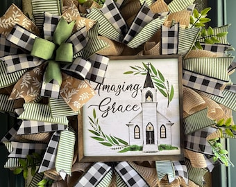 Könnte beinhalten: Ein dekorativer Kranz mit einem Holzschild mit der Aufschrift "Amazing Grace". Der Kranz besteht aus einer Mischung aus grünen, schwarz-weißen Büffelkaros und Jutabändern. Oben befindet sich eine grüne Schleife, und das Schild zeigt eine Kirche.