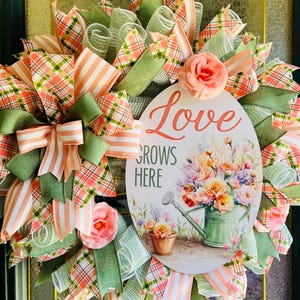 May include: A green, pink, and white wreath with a sign that reads "Love Grows Here". The sign features a watercolor illustration of a watering can with flowers.