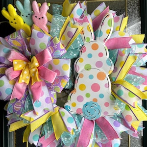 May include: A colorful Easter wreath with a bunny silhouette and three bunny figures. The wreath features ribbons in pink, yellow, and blue with polka dots and Easter egg patterns. A decorative spring wreath for the front door.