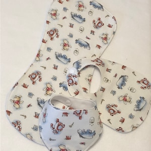 Op de afbeelding: Drie baby-slabbetjes met Winnie de Poeh-thema. Twee zijn wit met een Winnie de Poeh- en Biggetje-print en &eacute;&eacute;n is een wit bandana-slabbetje met dezelfde print.