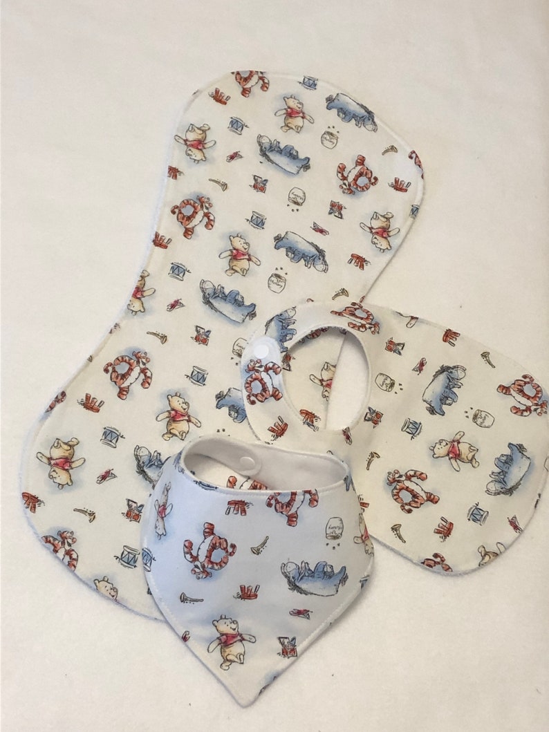 Op de afbeelding: Drie baby-slabbetjes met Winnie de Poeh-thema. Twee zijn wit met een Winnie de Poeh- en Biggetje-print en &eacute;&eacute;n is een wit bandana-slabbetje met dezelfde print.