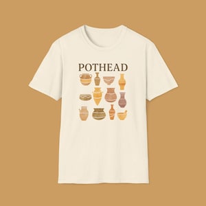Può includere: T-shirt color crema con la scritta "POTHEAD" sopra una collezione di illustrazioni di ceramiche in stile acquerello nei toni del marrone, giallo e arancione. La maglietta ha un girocollo classico e maniche corte.