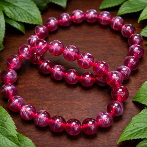 May include: Two ruby-colored beaded bracelets. Each bracelet is composed of round, polished beads with a star-like pattern. The bracelets are arranged on a dark wooden surface, with green leaves in the background.