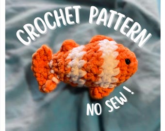 May include: A crocheted orange and white fish toy with black eyes. The text "CROCHET PATTERN" and "NO SEW!" are written in white cursive. The fish is made of a soft, textured yarn.
