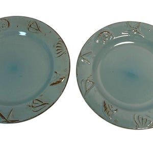 May include: Two light blue ceramic plates with embossed seashell designs around the rims. The plates have a slightly distressed look with brown accents, creating a coastal or nautical theme. The plates are round.