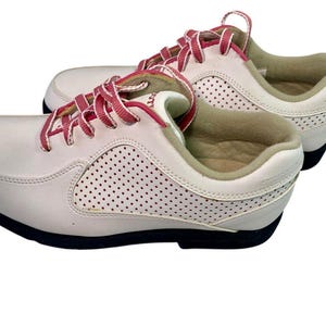 May include: Pair of white athletic shoes with pink laces. The shoes have a perforated design on the sides and a black sole. The interior lining is a light tan color.
