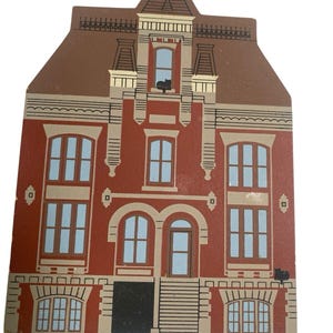 May include: A flat, decorative wooden cutout shaped like a multi-story house with a brown roof, red brick-colored walls, and light tan trim. The house features light blue windows, a central front door, and two small black cat silhouettes.