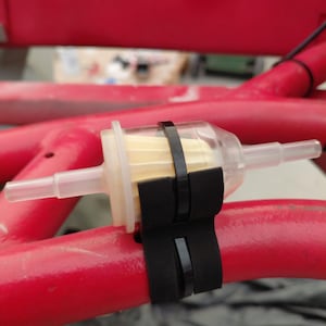 Peut inclure: Un filtre à carburant transparent avec un élément filtrant jaune et des tubes d'entrée et de sortie en plastique transparent. Le filtre est fixé à un cadre métallique rouge avec deux sangles noires.