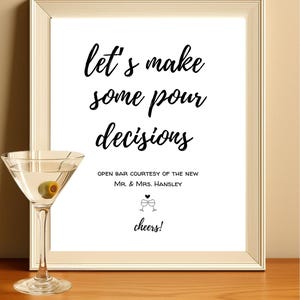 May include: A framed sign with the text "let's make some pour decisions" in black script. Below the text is "OPEN BAR COURTESY OF THE NEW MR. & MRS. HANSLEY" and "cheers!". A martini glass with an olive sits to the left of the sign.