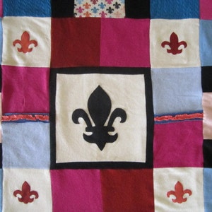 May include: A patchwork quilt with a variety of colors and patterns, including pink, red, blue, and white. The quilt is made up of squares and rectangles, and each square has a different design. The quilt is decorated with a fleur-de-lis design in the center.