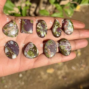 Könnte beinhalten: Neun polierte Edelsteine in verschiedenen Formen, darunter oval, tropfenförmig und rechteckig. Die Steine zeigen eine Mischung aus Farben, mit dominanten Farbtönen von Grün, Lila und Creme. Die Edelsteine werden auf einer Hand präsentiert.