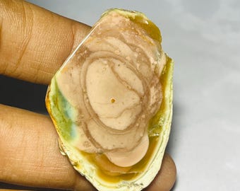 May include: A polished, irregularly shaped gemstone with a layered pattern of beige, brown, and yellow hues. The stone has a smooth, glossy surface and a natural, organic appearance. The gemstone is held in a hand.