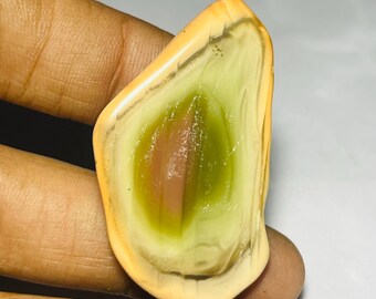 May include: A polished, oval-shaped gemstone with a unique pattern. The stone features a central area of green and pink hues, surrounded by layers of cream and tan colors. The gemstone is held in a hand, showcasing its natural beauty.