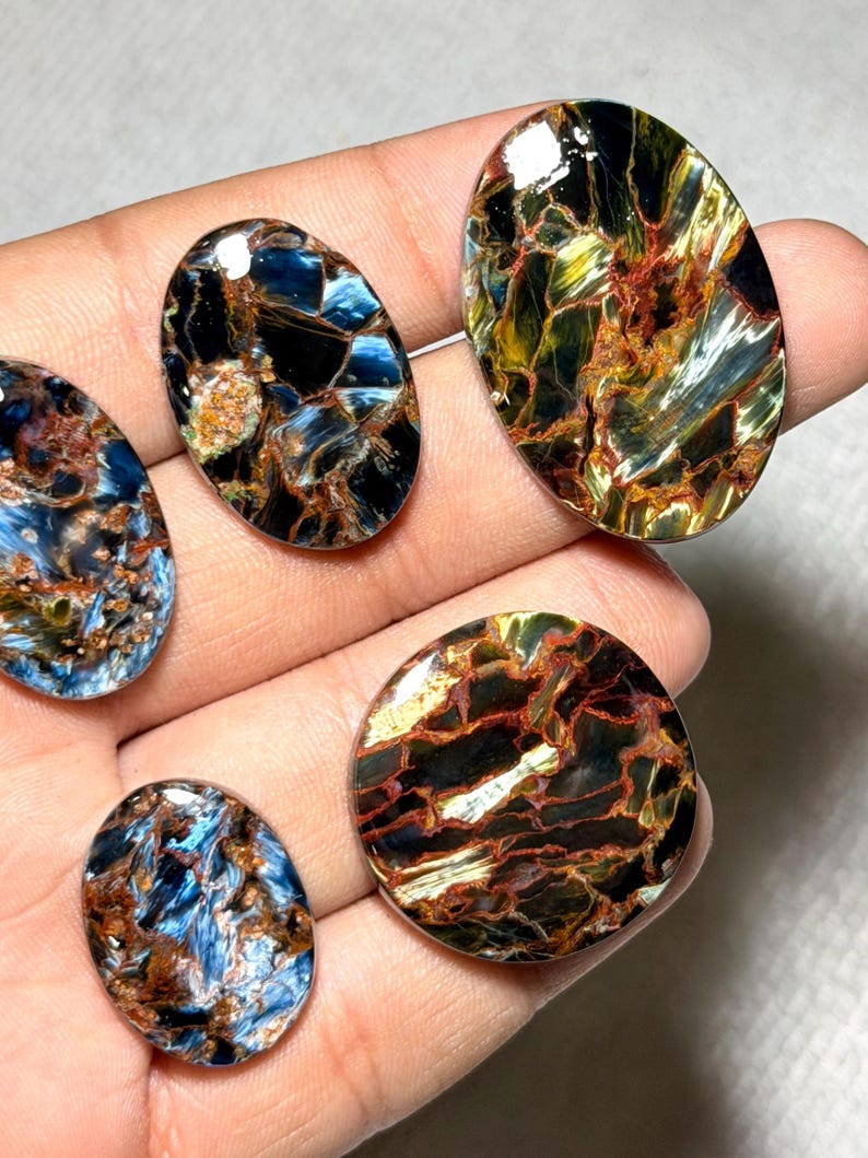 K&ouml;nnte beinhalten: Eine Sammlung polierter Pietersite-Edelsteine in verschiedenen ovalen und runden Formen. Die Steine zeigen eine Mischung aus blauen, braunen, goldenen und schwarzen Farben mit einzigartigen, wirbelnden Mustern. Die Edelsteine werden in einer Hand gehalten.