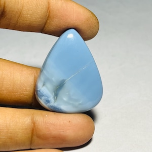 Pode incluir: Uma pedra preciosa polida em forma de gota, em tons de azul claro e branco. A pedra tem uma superfície lisa e brilhante com um padrão sutil e marmoreado. Uma linha fina e clara atravessa a superfície. A pedra preciosa é segurada entre dois dedos.