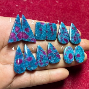 Könnte beinhalten: Eine Sammlung polierter Edelsteine in verschiedenen Formen, darunter Dreiecke, Quadrate und Tropfen. Die Steine weisen eine leuchtende türkisblaue Grundfarbe mit unregelmäßigen Flecken von tiefem Magenta auf. Die Edelsteine werden vor rotem Hintergrund präsentiert.