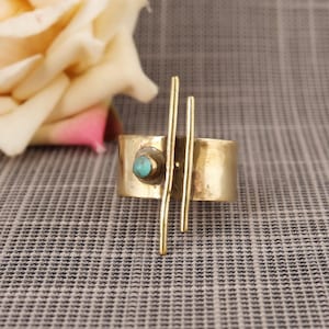 May include: A wide, brass-coloured ring with a turquoise stone and vertical gold bars. The ring has a band-like design with a small, round turquoise stone set on one side. Two vertical gold bars are attached to the ring.