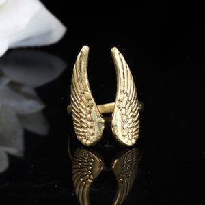 Op de afbeelding: Een goudkleurige ring met een ontwerp van twee open vleugels. De vleugels zijn gedetailleerd met veermotieven en bevinden zich aan weerszijden van de ringband. De ring staat tegen een zwarte achtergrond, wat de metaalglans benadrukt.