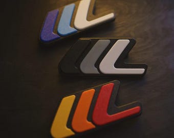 May include: Three automotive emblem badges in a close-up shot. The badges feature a stylized "W" and "L" design in various color combinations: purple, blue, and white; black, gray, and white; and yellow, orange, and red.