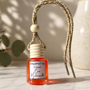 May include: A small, square glass bottle filled with orange liquid, featuring a white label that reads Inspired by Chanel Coco Mademoiselle. The bottle has a ribbed wooden cap and a braided beige and gold cord with a wooden bead for hanging.