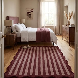 May include: A rectangular area rug featuring a scalloped edge and a vertical striped pattern in alternating shades of deep burgundy and muted mauve. The rug is positioned on a light-toned wooden floor in a bedroom setting with a bed and furniture.