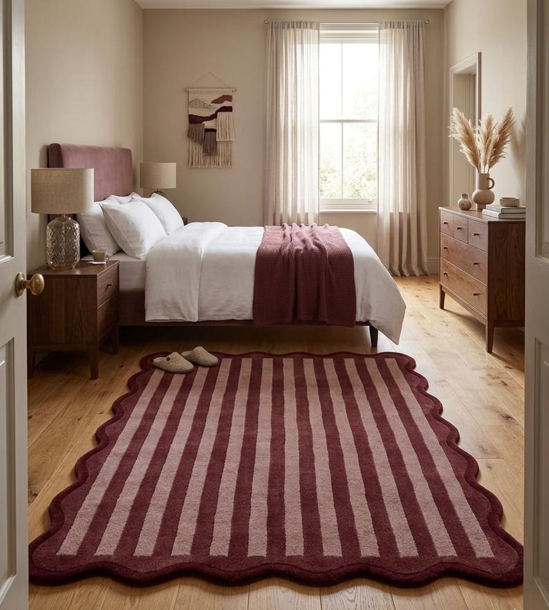 May include: A rectangular area rug featuring a scalloped edge and a vertical striped pattern in alternating shades of deep burgundy and muted mauve. The rug is positioned on a light-toned wooden floor in a bedroom setting with a bed and furniture.