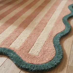 May include: A rectangular rug featuring alternating vertical stripes in soft pink and cream. The rug has a distinct scalloped edge finished with a thick, textured border in a deep teal green color, creating a wavy perimeter around the entire piece.