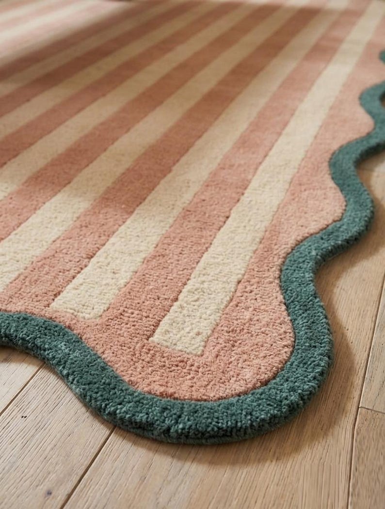 May include: A rectangular rug featuring alternating vertical stripes in soft pink and cream. The rug has a distinct scalloped edge finished with a thick, textured border in a deep teal green color, creating a wavy perimeter around the entire piece.