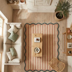 May include: A rectangular area rug featuring vertical stripes in muted coral and cream, finished with a dark teal scalloped border. The rug is positioned on a light wood floor in a living room setting with a sofa, coffee table, and chair.