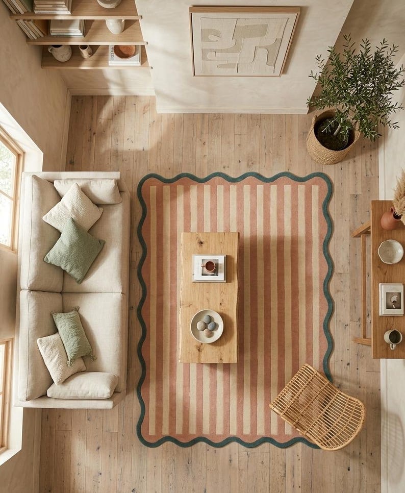 May include: A rectangular area rug featuring vertical stripes in muted coral and cream, finished with a dark teal scalloped border. The rug is positioned on a light wood floor in a living room setting with a sofa, coffee table, and chair.