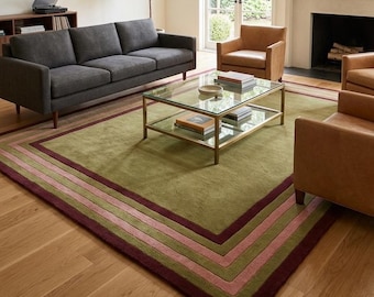 May include: A large rectangular area rug featuring a solid olive green center field framed by concentric borders in shades of muted pink and deep burgundy. The rug is positioned on a light-toned wooden floor beneath a glass-topped coffee table.