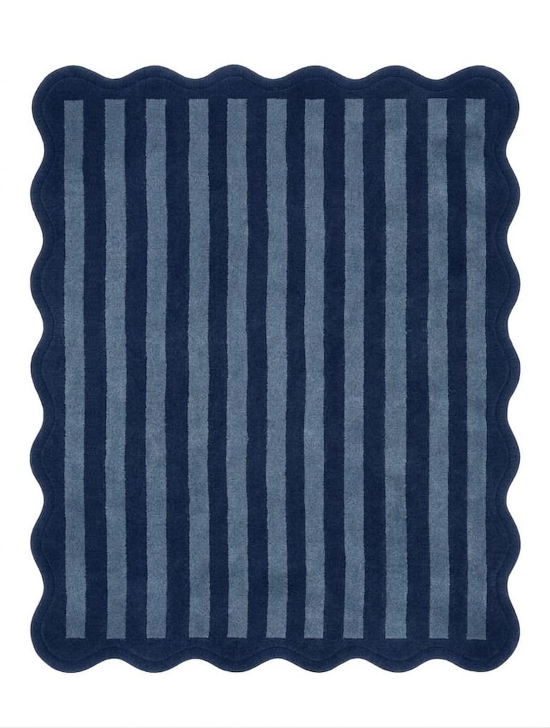 May include: A rectangular bath mat featuring a scalloped edge and a vertical striped pattern in alternating shades of dark navy blue and muted slate blue. The textile surface has a consistent, soft-textured pile throughout the entire design.