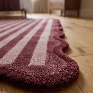 May include: A rectangular rug featuring a deep burgundy and light pink striped pattern. The rug has a distinct scalloped edge design along the side, with a thick, tufted texture that covers the entire surface of the floor covering.