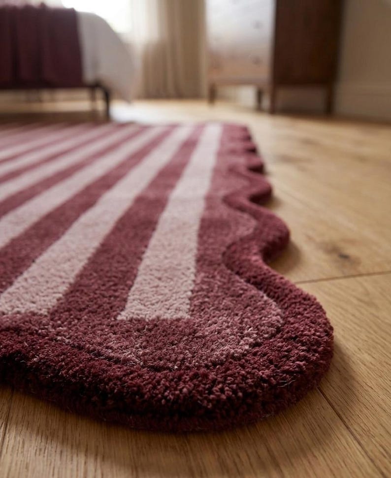 May include: A rectangular rug featuring a deep burgundy and light pink striped pattern. The rug has a distinct scalloped edge design along the side, with a thick, tufted texture that covers the entire surface of the floor covering.