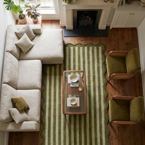 May include: A rectangular area rug featuring a scalloped edge and a vertical striped pattern in alternating shades of olive green and light sage. The rug is positioned on a dark wood floor, centered beneath a glass-topped coffee table.