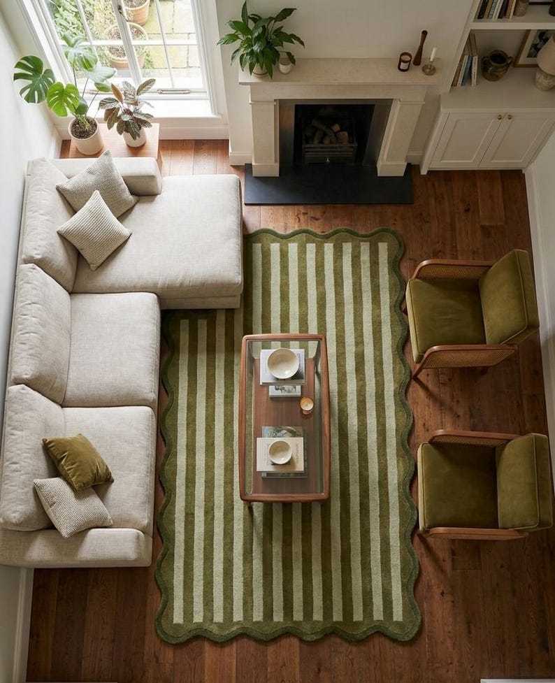 May include: A rectangular area rug featuring a scalloped edge and a vertical striped pattern in alternating shades of olive green and light sage. The rug is positioned on a dark wood floor, centered beneath a glass-topped coffee table.