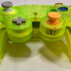 Könnte beinhalten: Ein transparenter neongrüner Videospiel-Controller mit grauen und orangefarbenen Knöpfen. Der Controller hat ein grünes Kabel. Die Tasten umfassen ein Steuerkreuz, Analog-Sticks und Aktionstasten. Der Controller ist für Spiele konzipiert.