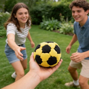 Op de afbeelding: Een geel en zwarte voetbal wordt in de hand van een persoon gehouden, met twee mensen op de achtergrond. De bal heeft een klassiek voetbalontwerp met zwarte vijfhoeken en gele zeshoeken. De achtergrond is een groen gazon.