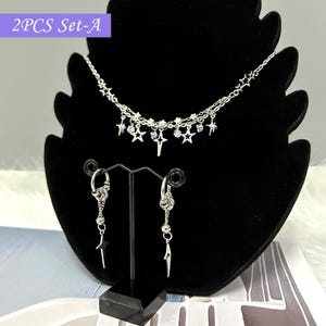 Op de afbeelding: Een zilverkleurige sieradenset gepresenteerd op een zwarte standaard. De set bevat een gelaagde ketting met ster- en kristallen bedels, en bijpassende oorringen met bungelende ster- en geometrische accenten. De tekst "2PCS Set-A" staat linksboven.